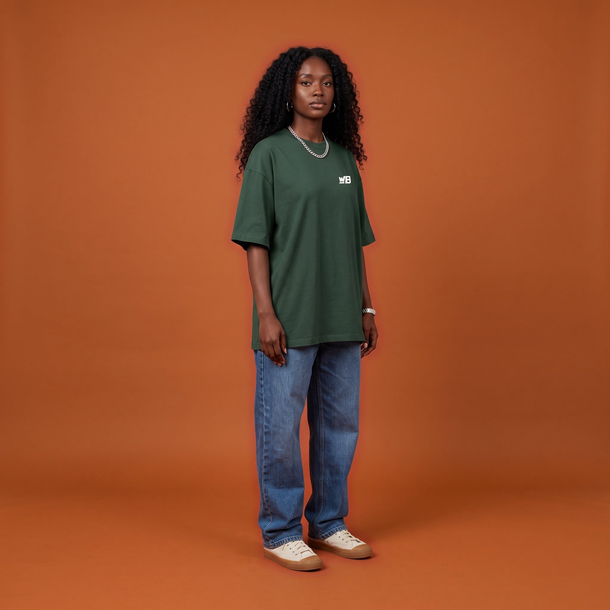 Blackness Sankofa Queen Tee, deep forest green, worn by a model — front view with WB chest logo, terracotta backdrop