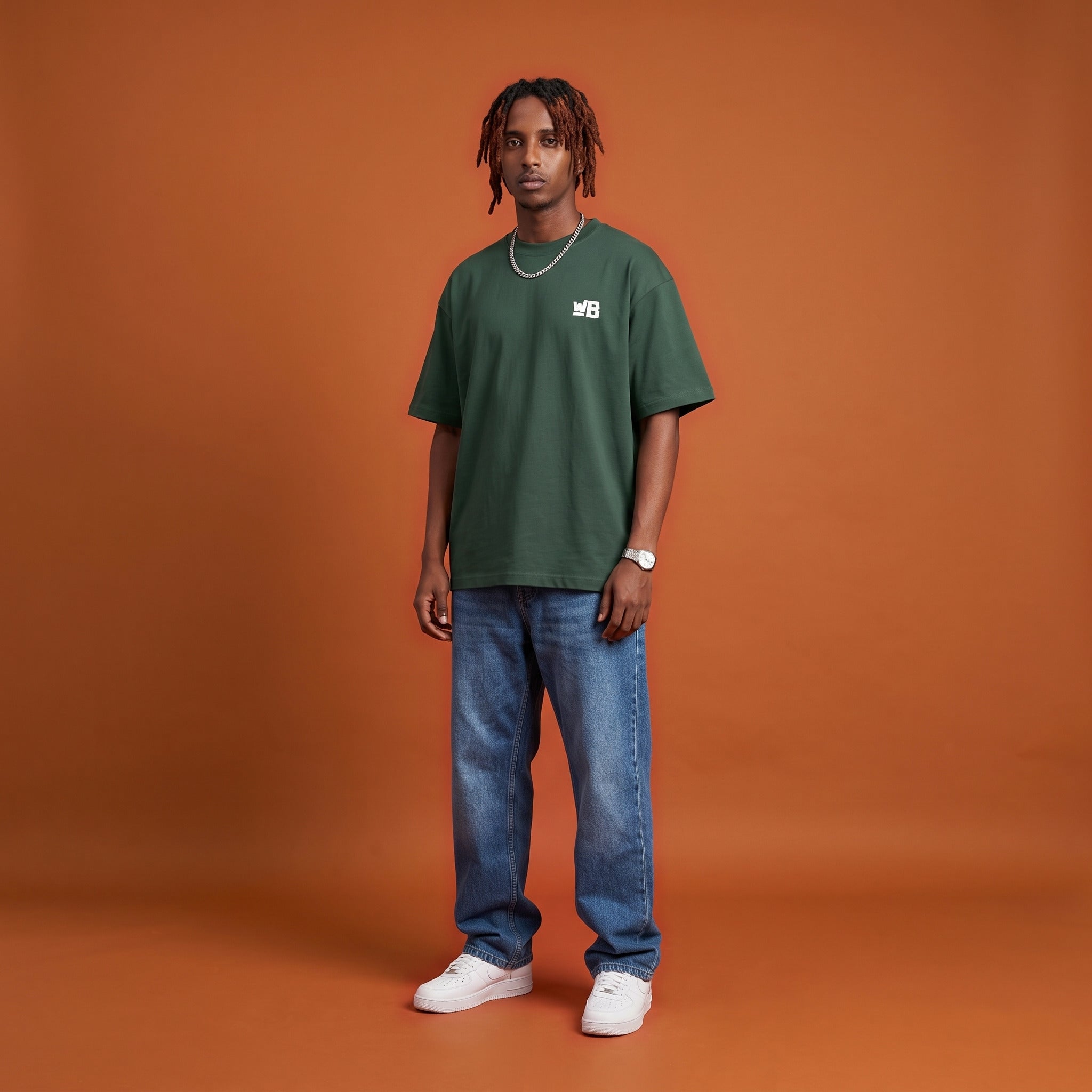Blackness Sankofa King Tee, deep forest green, worn by a model — front view with WB chest logo, terracotta backdrop