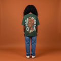Blackness Sankofa Queen Tee, deep forest green, worn by a model — back view, Sankofa headwrap portrait print, terracotta backdrop