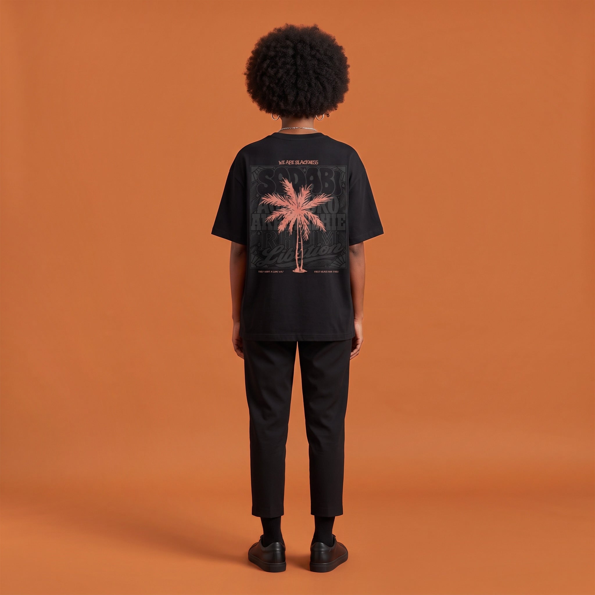 Blackness Libation Tee, black, worn by a model — back view, palm tree Libation print, terracotta-orange studio backdrop