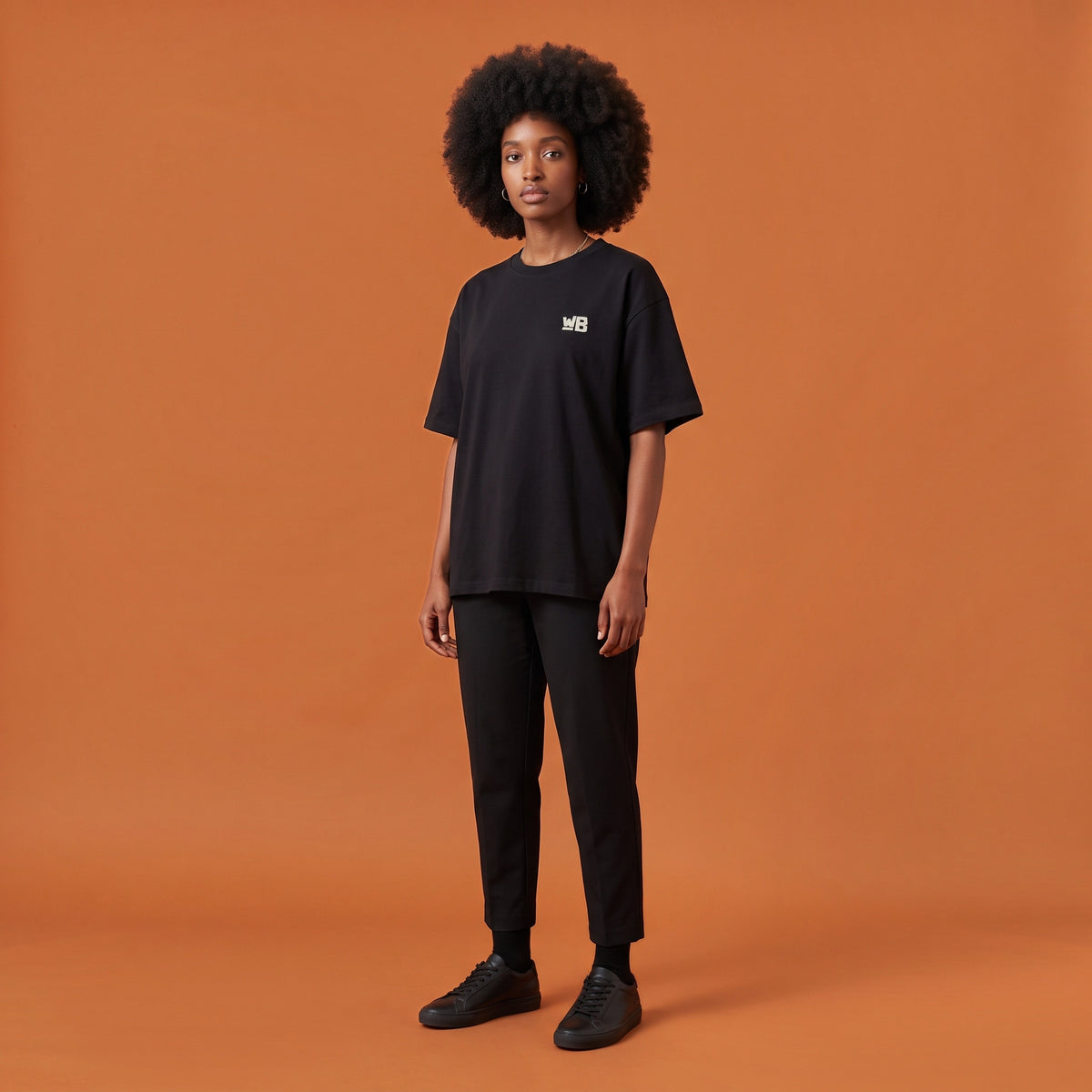 Blackness Libation Tee, black, worn by a model — front view with WB chest logo, terracotta-orange studio backdrop