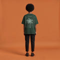 Blackness Libation Tee, forest green, worn by a model — back view, palm tree Libation print, terracotta-orange studio backdrop