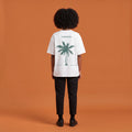 Blackness Libation Tee, white, worn by a model — back view, palm tree Libation print, terracotta-orange studio backdrop