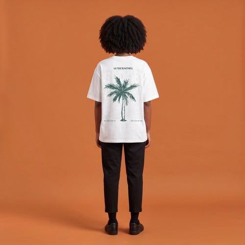 Blackness Libation Tee, white, worn by a model — back view, palm tree Libation print, terracotta-orange studio backdrop