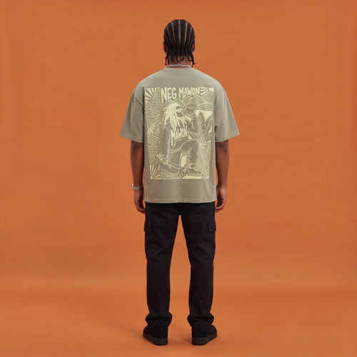 Blackness Mawon Tee, brown khaki, worn by a model — back view, Neg Mawon conch-blower block print, terracotta-orange studio backdrop