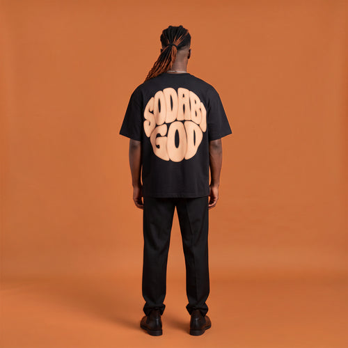 Blackness Sodabi Tee, black, worn by a model — back view, SODABI GOD bubble lettering print, terracotta-orange studio backdrop