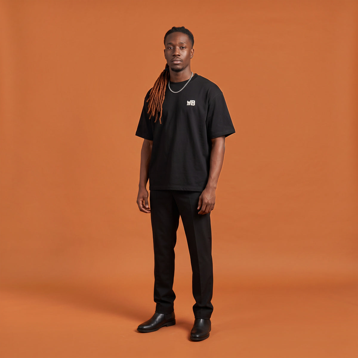 Blackness Sodabi Tee, black, worn by a model — front view with WB chest logo, terracotta-orange studio backdrop