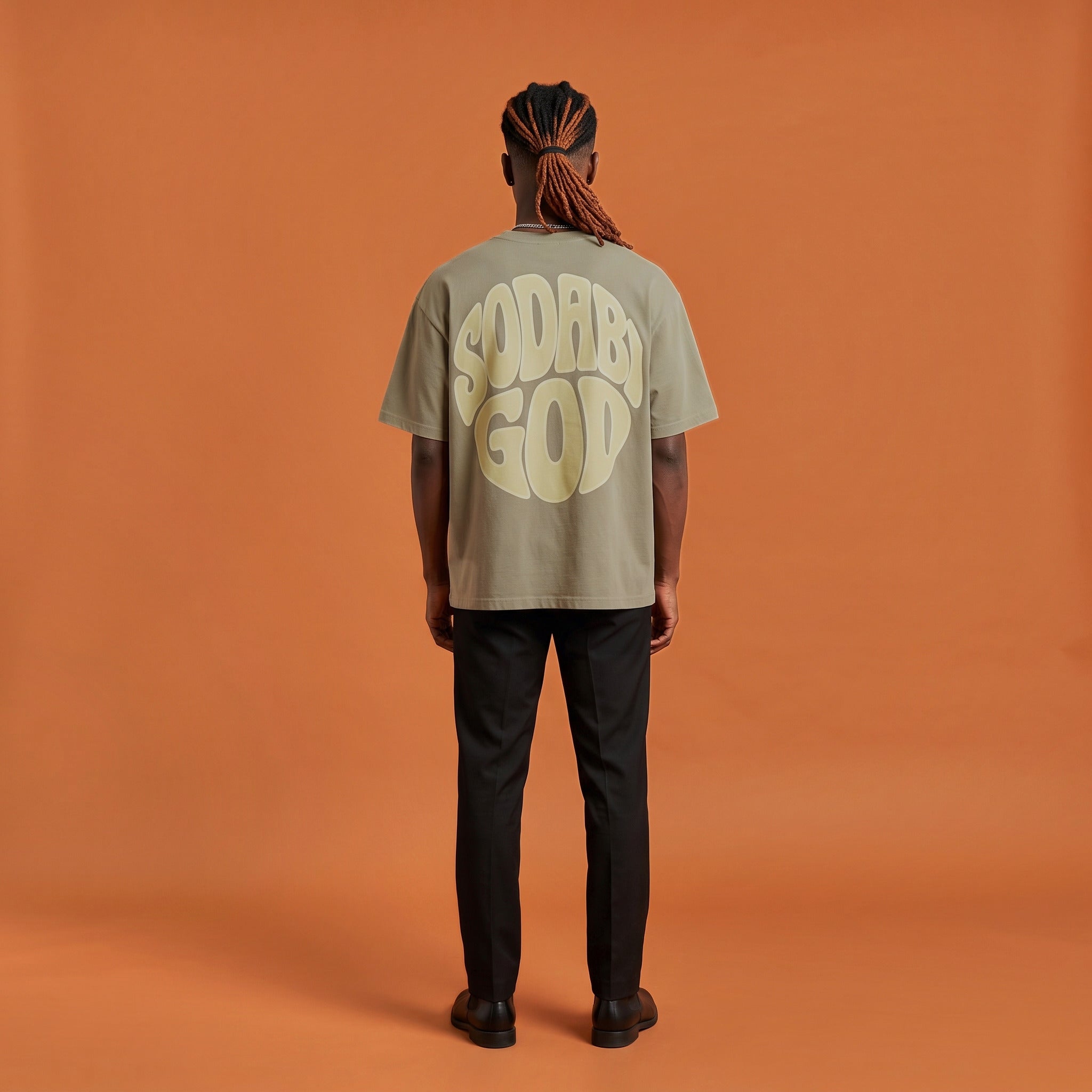Blackness Sodabi Tee, brown khaki, worn by a model — back view, SODABI GOD bubble lettering print, terracotta-orange studio backdrop