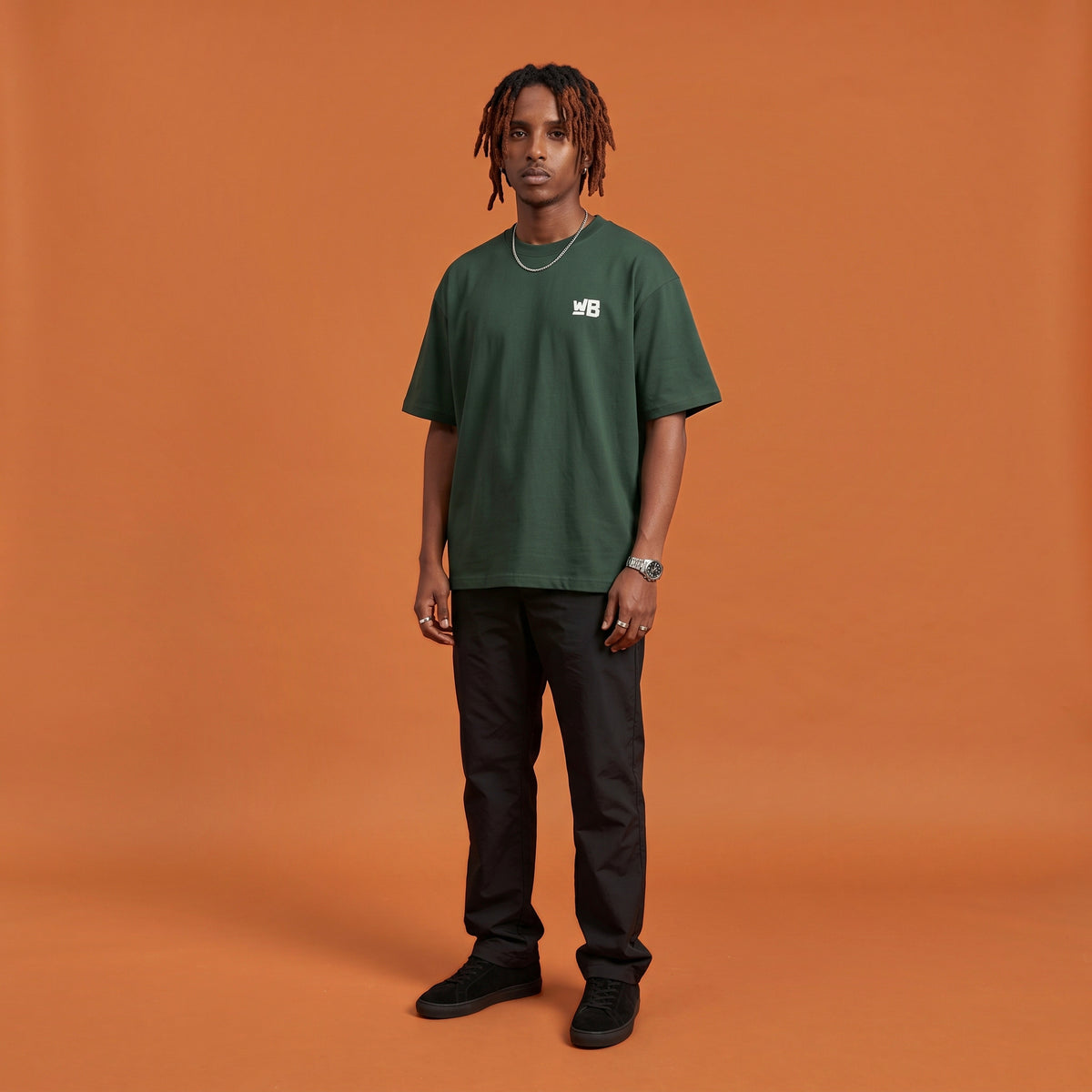 Blackness Sodabi Tee, forest green, worn by a model — front view with WB chest logo, terracotta-orange studio backdrop