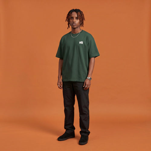 Blackness Sodabi Tee, forest green, worn by a model — front view with WB chest logo, terracotta-orange studio backdrop