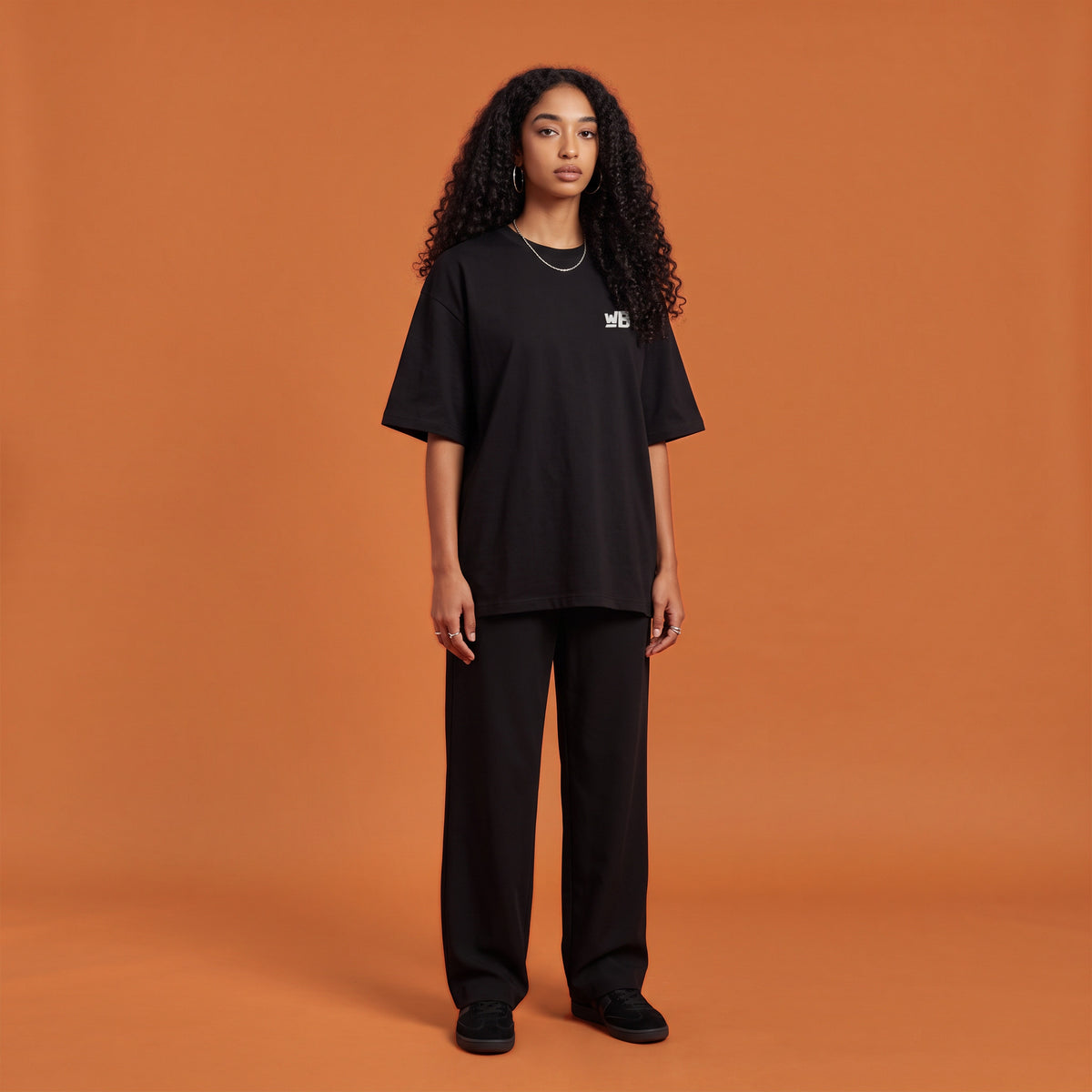 Blackness Solitude Tee, black, worn by a model — front view with WB chest logo, terracotta-orange studio backdrop