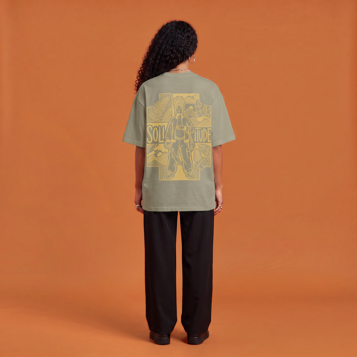 Blackness Solitude Tee, brown khaki, worn by a model — back view, Solitude portrait block print, terracotta-orange studio backdrop