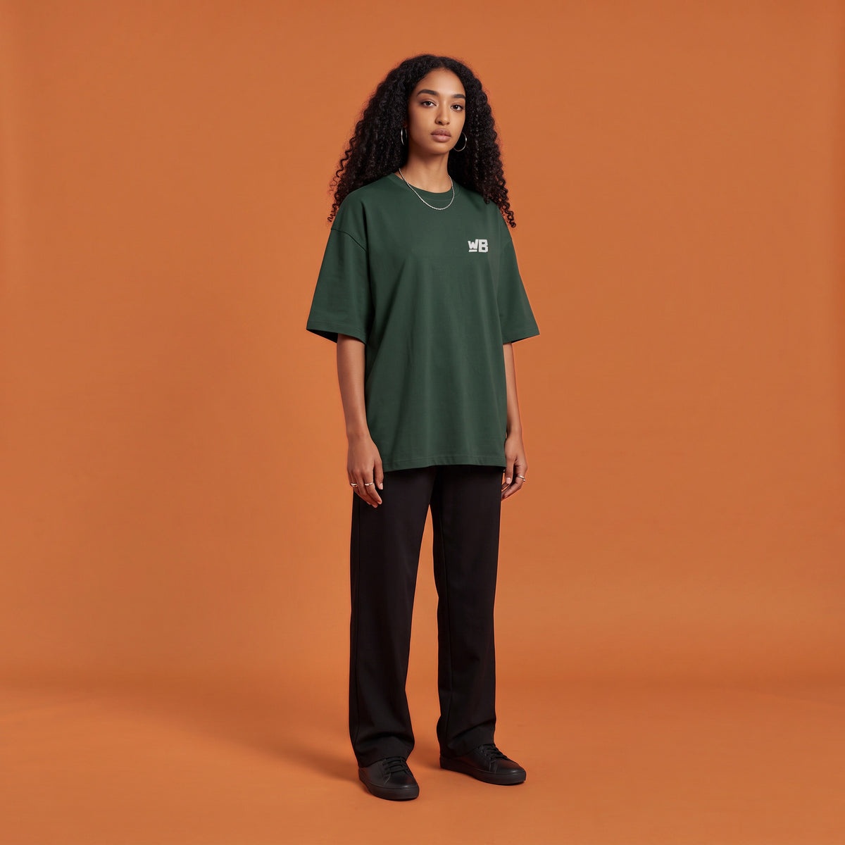 Blackness Solitude Tee, forest green, worn by a model — front view with WB chest logo, terracotta-orange studio backdrop