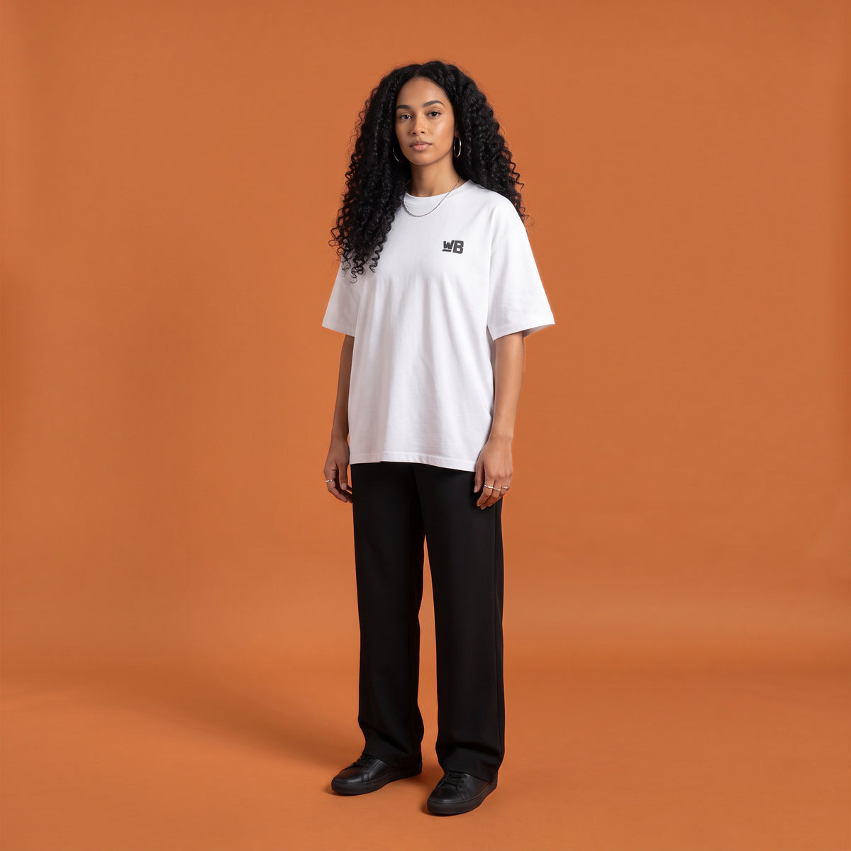 Blackness Solitude Tee, white, worn by a model — front view with WB chest logo, terracotta-orange studio backdrop