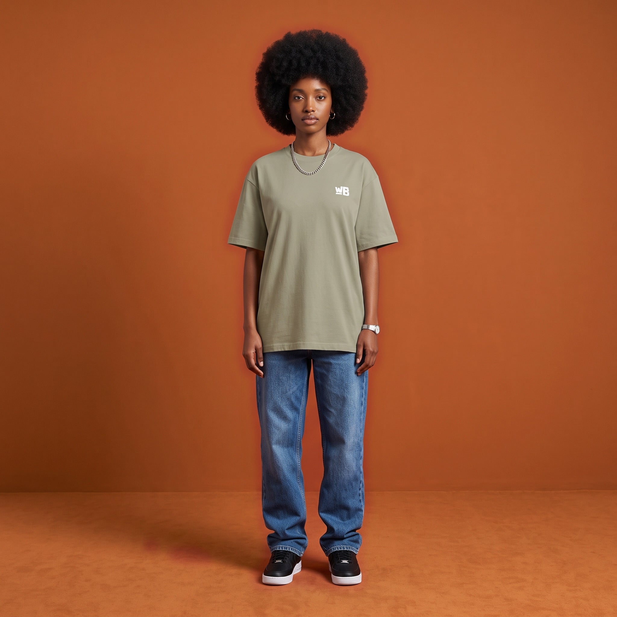 Blackness Sankofa Queen Tee, brown khaki, worn by a model — front view with WB chest logo, burnt-orange terracotta studio backdrop