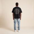 Blackness Emancipation Tee Black worn by model, back view showing the spiral Emancipate Yourself print on beige studio backdrop