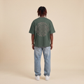 Blackness Emancipation Tee Cactus Green worn by model, back view showing the spiral Emancipate Yourself print on beige studio backdrop