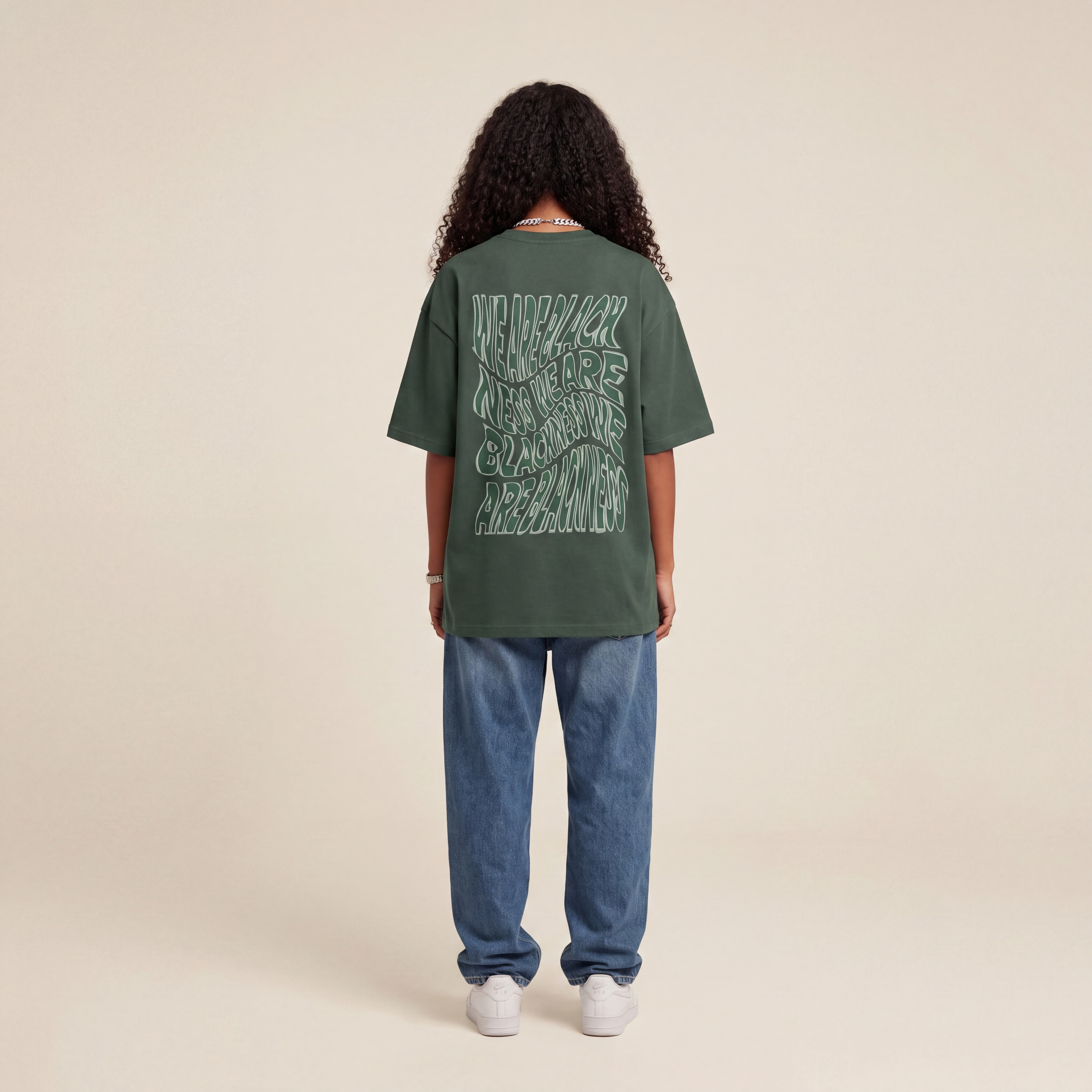 Blackness Honor & Embrace Tee Cactus Green worn by model, back view showing the We Are Blackness wavy print on beige studio backdrop