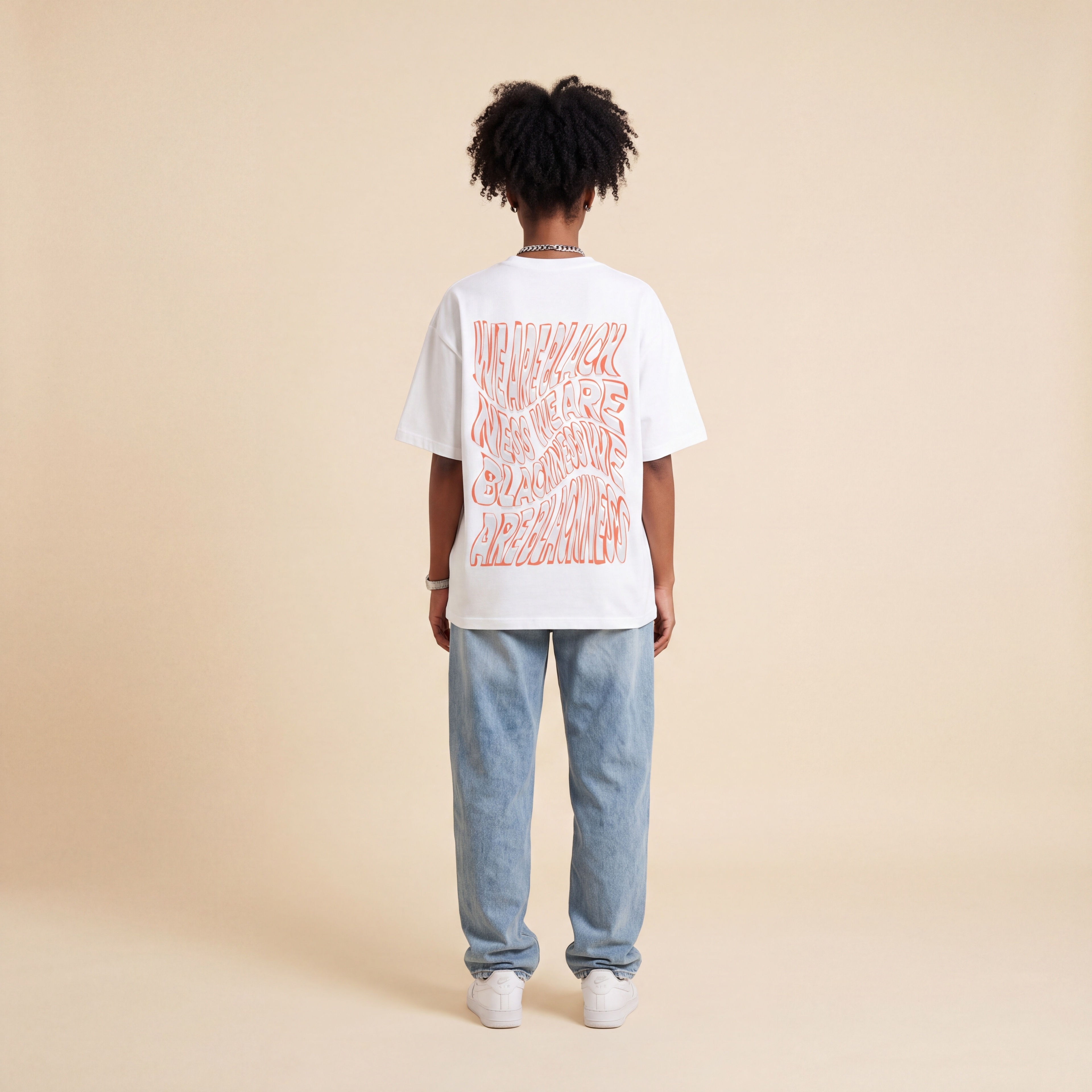 Blackness Honor & Embrace Tee White worn by model, back view showing the We Are Blackness wavy print on beige studio backdrop