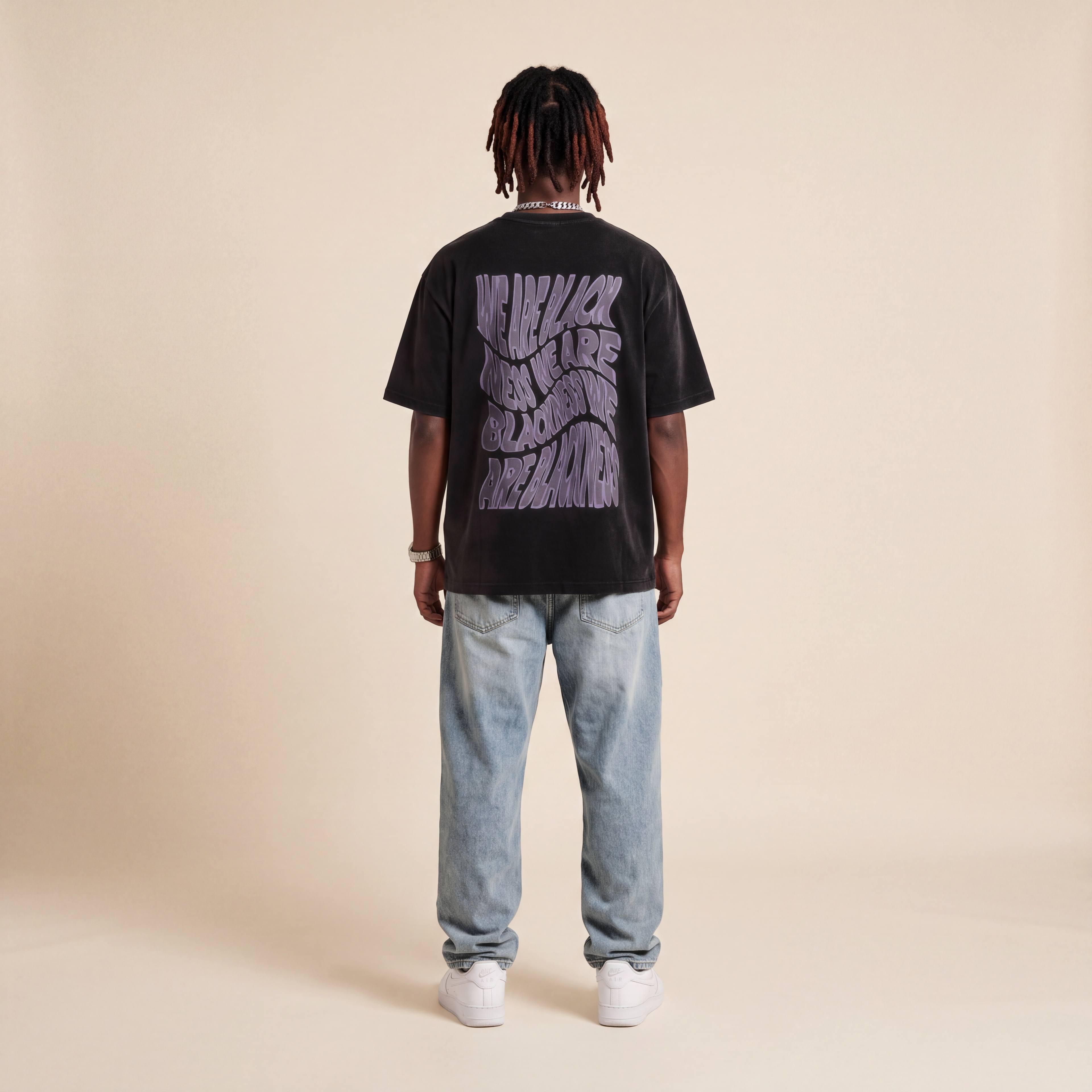 Blackness Honor & Embrace Tee Black worn by model, back view showing the We Are Blackness wavy print on beige studio backdrop